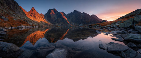 A mountain range is reflected in the tranquil waters of a lake. The scene displays the colors of a sunset, with orange and blue hues dominating the sky. The foreground features rocks. This image may be useful for travel, environment, or scenic design projects.の素材