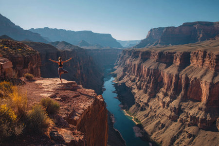 A person stands atop a canyon, arms raised, against a backdrop of towering rock formations and a river. Warm light bathes the scene, highlighting the textures of the cliffs and the vastness of the landscape. This image is suitable for travel articles, environmental campaigns, and adventure-related content.の素材