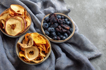 An overhead view presents assorted dried fruits in small ceramic bowls. The composition features a variety of textures and colors. The setup is placed on a textured grey textile. It could be suitable for illustrating healthy eating, snacks, or culinary articles.の素材