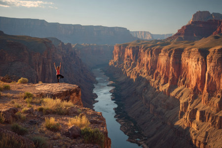 A person performs yoga on a cliff edge. The warm tones of the rock contrast with the blue river flowing through the canyon. The image features a wide composition with natural lighting, suitable for projects related to health, wellness, and travel. It could be utilized for various editorial or commercial purposes.の素材