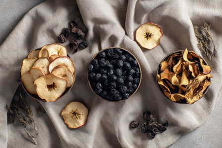 This overhead shot presents an arrangement of dried fruits, including apple slices and other varieties, displayed in small bowls. The composition emphasizes textures and forms. The soft lighting enhances the natural colors against a neutral-toned linen cloth. This image could be used in culinary projects, healthy eating promotions, or food-related editorial content.の素材
