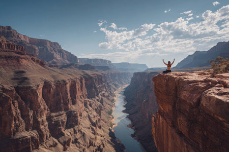 A person sits atop a rock formation, arms raised, with a vast canyon stretching out before them. The scene features a clear blue sky, a winding river, and the earthy tones of the rock formations. This composition could be used for editorial features or commercial purposes.の素材