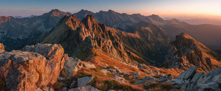An expansive view showcases a rugged mountain range at dawn. The composition displays sharp peaks, layered valleys, and rocky terrain in varying shades of orange and brown. The warm sunlight creates a dramatic contrast, suggesting a serene natural environment, useful for travel or outdoor themes.の素材