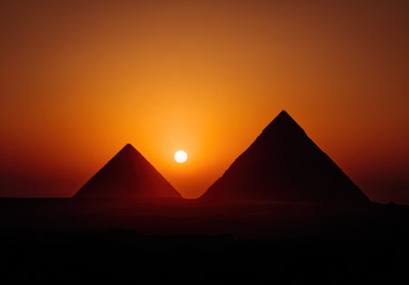 Two ancient pyramids are silhouetted against a vibrant, orange sunset. The composition features a strong contrast between the dark, geometric shapes of the structures and the fiery sky. This image could be used for travel promotions, historical publications, or educational materials, providing a sense of wonder.の素材