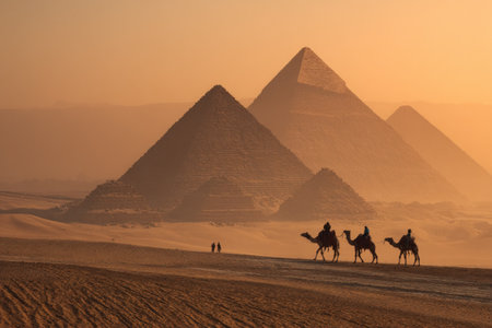 The image showcases the iconic pyramids with silhouettes of camels and riders in the foreground. The warm tones of the setting sun create a hazy, golden atmosphere. The composition emphasizes the scale of the structures against the vast desert landscape. This scene could be used for travel, historical, and tourism-related content.の素材