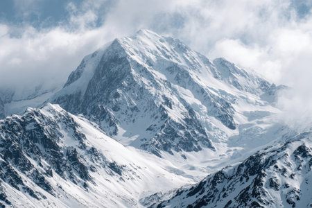 An aerial view captures a large snow-covered mountain peak surrounded by clouds. The image displays a stark white and blue color palette with textures of snow and rock. The composition highlights the peak against a bright, overcast sky, suitable for commercial and editorial applications.の素材