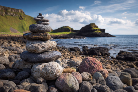 A stone cairn stands on a rocky shore, with a scenic coastal background. The image showcases various gray and brown stones, with a vibrant blue ocean and sky. The composition and lighting suggest outdoor daylight, perfect for illustrating themes of balance and environment in various commercial projects.の素材
