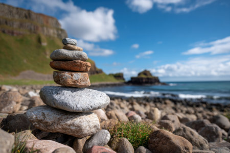 A carefully balanced stack of rocks dominates the foreground of a coastal scene. Various shades of brown and grey stones are precisely arranged. The background shows the sea, land, and a partly cloudy blue sky. This image could be used in editorial and commercial projects.の素材