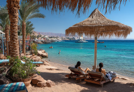 Two people relax on lounge chairs under a straw umbrella on a tropical beach. Palm trees and turquoise water provide the backdrop, with hints of a town and boats in the distance. Warm sunlight illuminates the scene, with potential applications for travel, leisure, or lifestyle imagery.の素材