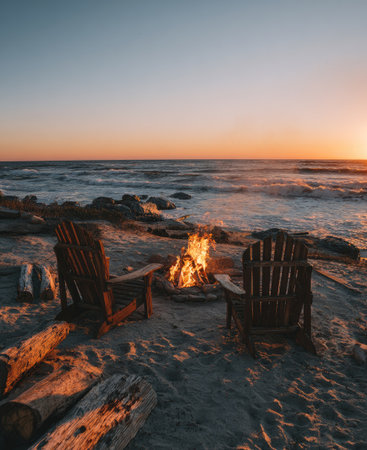 Two wooden chairs face a bonfire on a sandy beach. The image captures the warmth of a fire near the water with the sunset casting orange hues. The scene includes logs and the ocean waves under the evening sky, suitable for editorial or commercial imagery.の素材