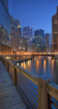 A twilight cityscape features tall buildings reflecting in tranquil water. The image displays a wooden walkway in the foreground, leading towards a panorama of skyscrapers. Warm lights from the buildings and their reflections create a contrast against the cool blue hues of the sky and water. Suitable for various commercial and editorial applications.の素材