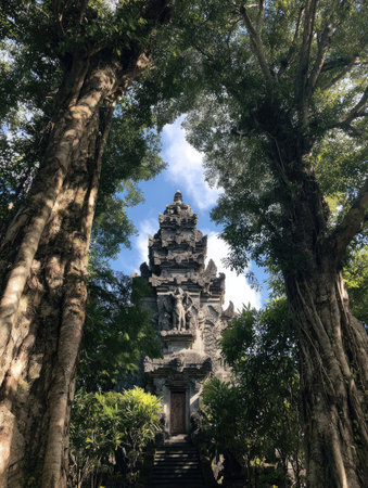 An upward perspective captures an ancient temple tower nestled amidst verdant trees under a vibrant blue sky. The composition emphasizes the structure's ornate details and the textured bark of the surrounding trees. This image may be suitable for various uses, including editorial articles, educational resources, and decorative applications.の素材