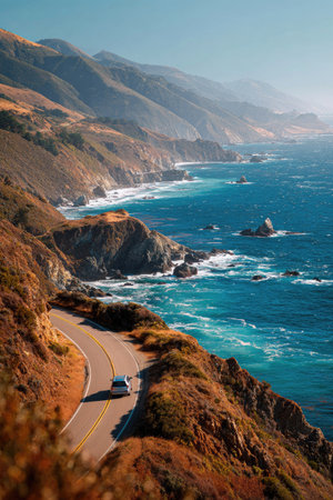 A vehicle travels along a winding coastal highway, showcasing a breathtaking landscape. The scene displays a deep blue ocean crashing against rocky shores, accompanied by a mountain range. The sunlight illuminates the natural colors and creates a sense of depth and tranquility. This image is suited for editorial and promotional content.の素材