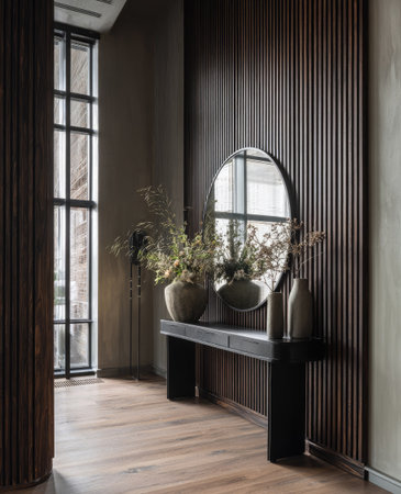 An interior shot reveals a stylish entryway featuring a dark console table adorned with vases and a large oval mirror. The space has wooden paneling and a large window, conveying a sense of sophistication and minimalist design. The image may be suitable for home decor, architectural, or lifestyle content.の素材