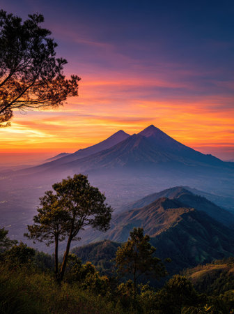 The image features mountain silhouettes under a vibrant sunset. The sky displays a gradient of colors from orange to purple. Trees are visible in the foreground, suggesting a mountainous environment. The scene could be utilized for landscape design, travel media, or editorial projects.の素材