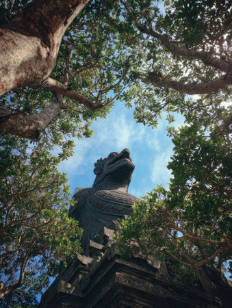 A stone sculpture is the focal point of this image, captured from a low angle. It is framed by a canopy of green trees set against a blue sky with some sunlight. The composition is a vertical shot. This photo could be used for various editorial and commercial purposes.の素材