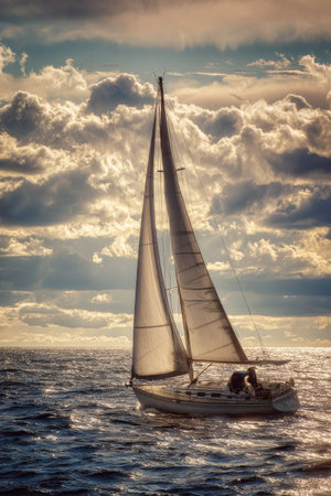 A sailboat glides across the ocean under a dramatic, cloudy sky. The image displays a warm color palette, with sunlight illuminating the water and the boat's sails. This picturesque scene could be used in various projects needing visual elements related to travel, nautical themes, or recreational activities.の素材