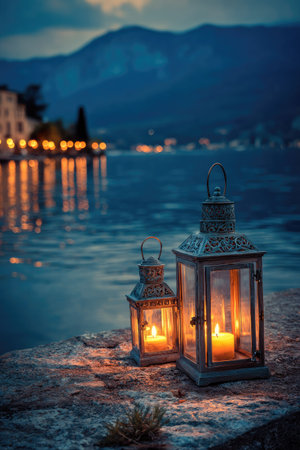 Two ornate lanterns with lit candles stand on a rocky surface, set against a tranquil lake scene. The composition features soft lighting, with a focus on warm colors emanating from the lanterns. The background includes a mountainous silhouette and the distant lights of a town. Suitable for atmospheric visuals or conceptual projects.の素材