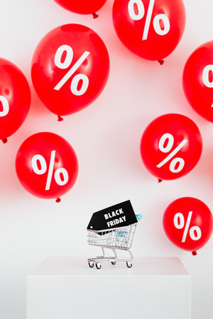 Shopping cart with percent sign on white cube and red balloons backgroundの素材