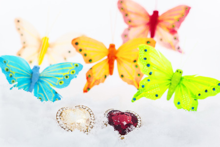 Two decorative hearts in the white snow on the background of multicolored decorative butterflies close up. The background is blurredの写真素材