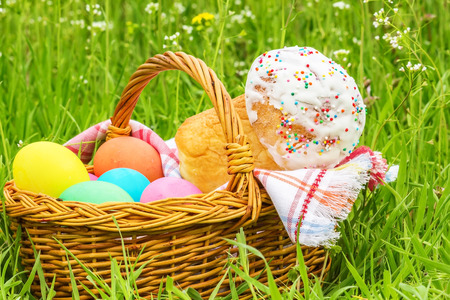 Wicker basket with painted eggs and Easter cake on the spring green grass, close up