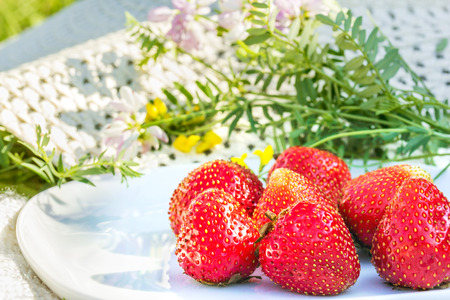 Red ripe strawberries on a white plate in the summer garden on a background of white hat and wildflowers. Selective focusの写真素材