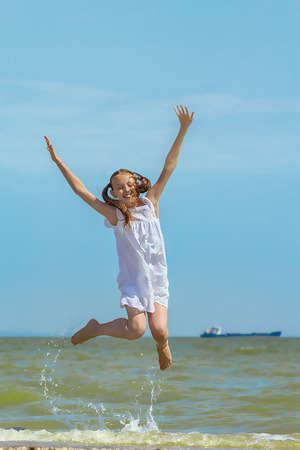 Happy girl in a white sundress  jumping over the water on the beach against the sea and blue sky with clouds on a hot summer dayの写真素材