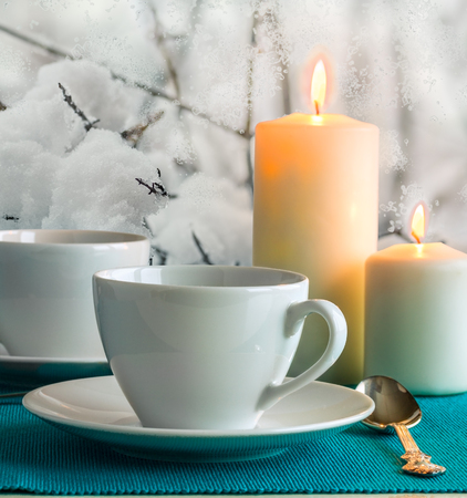 Two white cups and saucers, two burning candles and a tea spoon on a turquoise napkin on the background of snow-covered garden outside the window in winter evening, close-upの写真素材