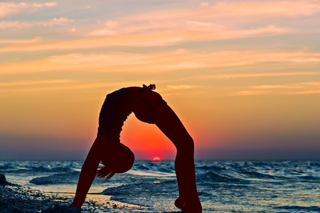 Girl-silhouette doing a gymnastic exercise on the beach against a backdrop of a beautiful sunsetの写真素材