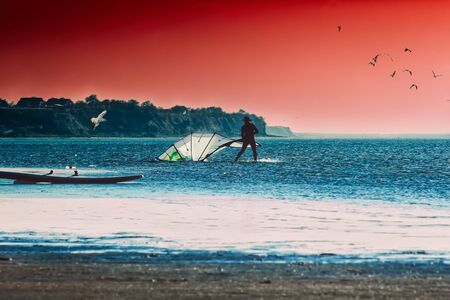 Silhouette of a surfer folds sail on the beach against the backdrop of the seascape and dramatic...