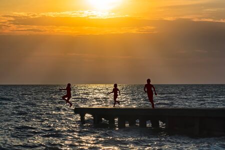 Silhouettes of children running on the breakwater and jumping into the sea against the orange sunsetの写真素材