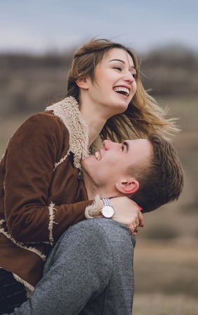 beautiful lovely couple in the park or field or track. man in coat and scarf with girl in coat. man hugs picked women up flirting laugh and look at each other. emotional soulful photo verticalの写真素材