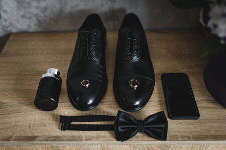 The groom is gathering in the morning. wooden shelf. Mens Black classic patent leather shoes. A black bow tie. Wedding details. Eau de toilette, black perfume, black mobile phoneの写真素材