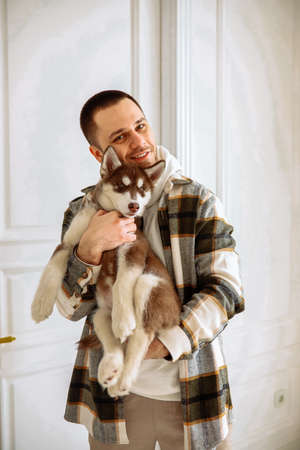 a young Caucasian man with dark hair in a plaid shirt plays with siberian husky dogs. The guy owner holding a husky puppy in a white roomの写真素材