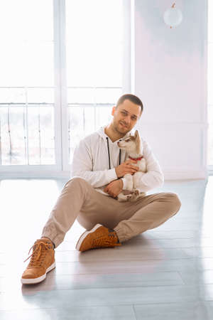 Attractive man with dark hair in a white sweater plays with siberian husky dogs. The man owner holding a husky puppy in a white room sitting on the floor.の写真素材