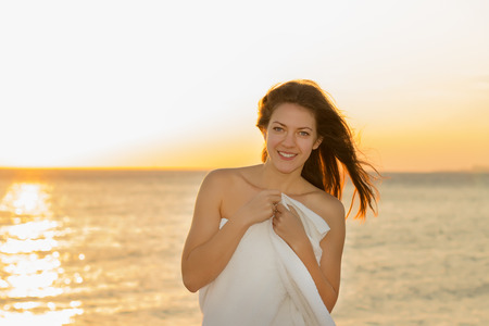 A girl wrapped in a towel smiling at sunset near the sea
