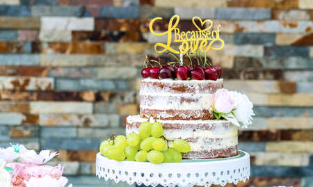 Multilayered wedding cake covered in white icing sugar and decorated with rosy red apples placed on a stand outside in the garden next to a flowering shrub. On top of the cake in gold is the word "love".の写真素材
