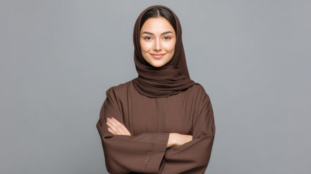 A young woman wearing a brown hijab and dress stands confidently with crossed arms. Her warm smile and serene expression exude strength, elegance, and cultural pride.の素材