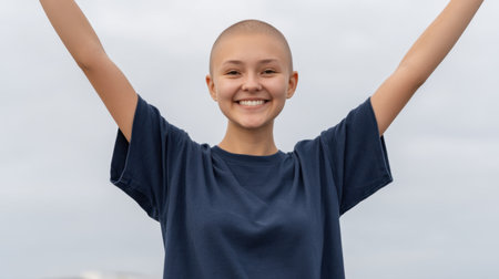 A young girl with a shaved head joyfully raises her arms outdoors, showcasing happiness and confidence against a cloudy backdrop in a casual blue t-shirt.の素材