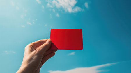 Close-up of hand holding a red card, blue sky backdrop, sunlight on the card, emphasizing the gestureの素材