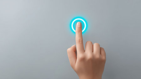 A close-up of a human finger gently touching a glowing blue circular button on a smooth gray surface, symbolizing interaction with modern technology and user engagement.の素材