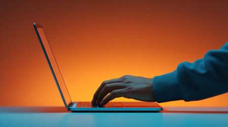 A close-up side view of a person's hand typing on a sleek laptop against a vibrant orange background, ideal for themes of technology, creativity, and productivity.の素材