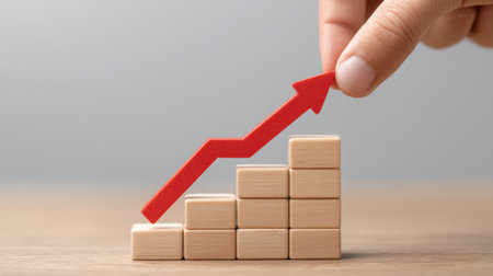 A hand is shown placing a red upward arrow on stacked wooden blocks, symbolizing growth and success in business or individual development strategies.の素材