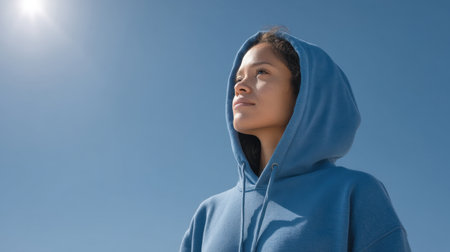 A young woman in a blue hoodie gazes upward, embracing the warmth of sunlight against a clear sky. This serene image embodies a sense of hope and positivity.の素材