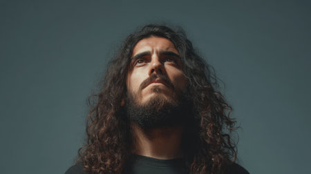 A striking portrait featuring a bearded man with long, curly hair looking confidently upwards. The neutral gray background highlights his features and calm demeanor.の素材