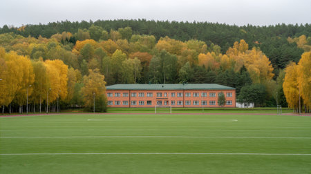 A modern school building stands in a picturesque setting, embraced by colorful autumn trees beside a lush green sports field under a cloudy sky.の素材