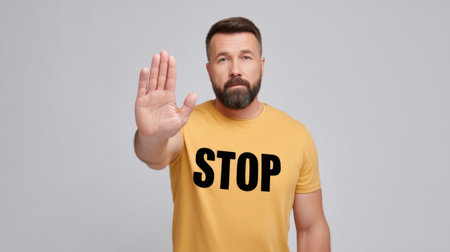 A man with a beard stands in a yellow shirt displaying a 'STOP' gesture with his raised hand, promoting the idea of pause and awareness in various contexts.の素材