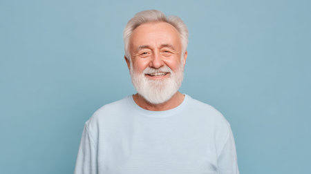 A joyful elderly man with a white beard and light blue sweater stands against a matching background, exuding positivity and warmth through his genuine smile.の素材