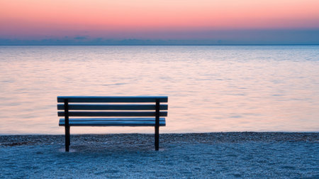 An empty bench on a quiet beach, overlooking the serene sea under a soft, pastel-colored sky at dusk.の素材
