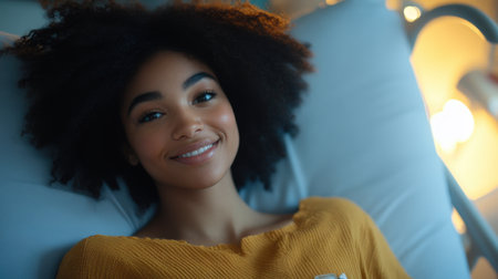 A joyful young woman with natural hair smiles while relaxing in bed, illuminated by soft light, creating a serene and cozy atmosphere filled with positivity and warmth.の素材